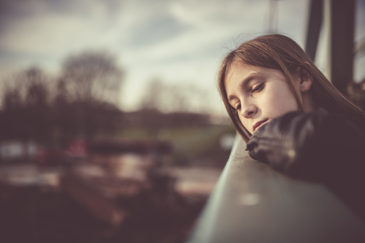 a young girl about 12 years old with long dark hair is leaning out and looking over a metal balcony. She is wearing a dark jacket. Her facial expression is sad and reflective to convey the effects of trauma bonding.