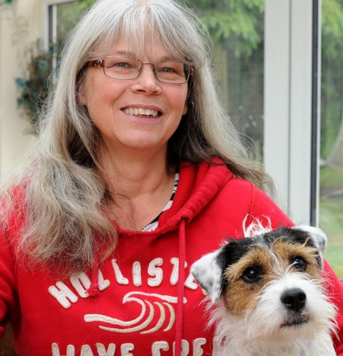 MBE awarded foster carer Judith with her dog. Judith is a white woman with long grey hair and a fringe. She is wearing glasses and is smiling. She is wearing a bright red hoodie sweatshirt with white writing on it. By her side is a small brown and white terrier dog.
